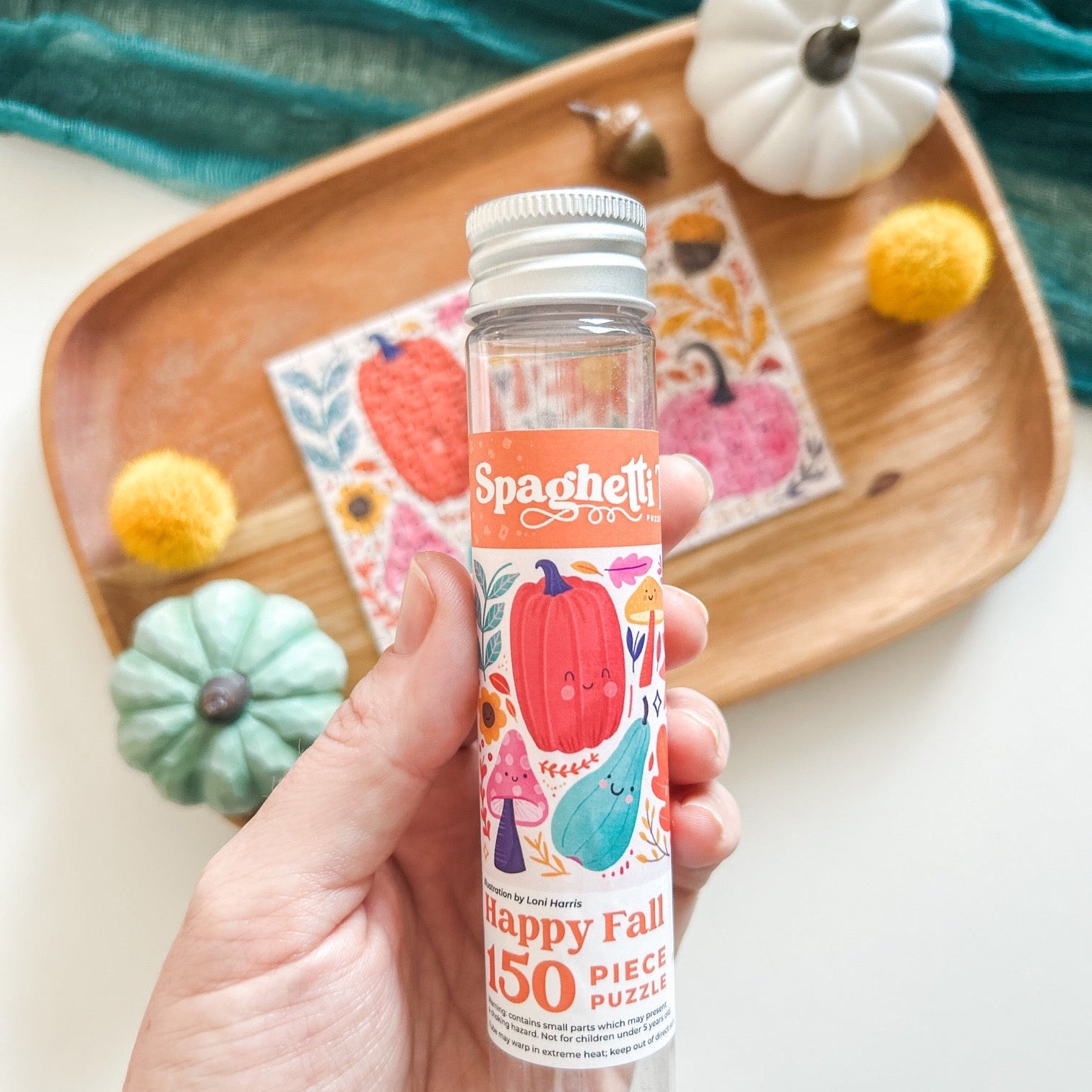 Happy Fall Puzzle in a small plastic tube, pictured above a wooden tray with tiny pumpkins and a teal gauze table runner.