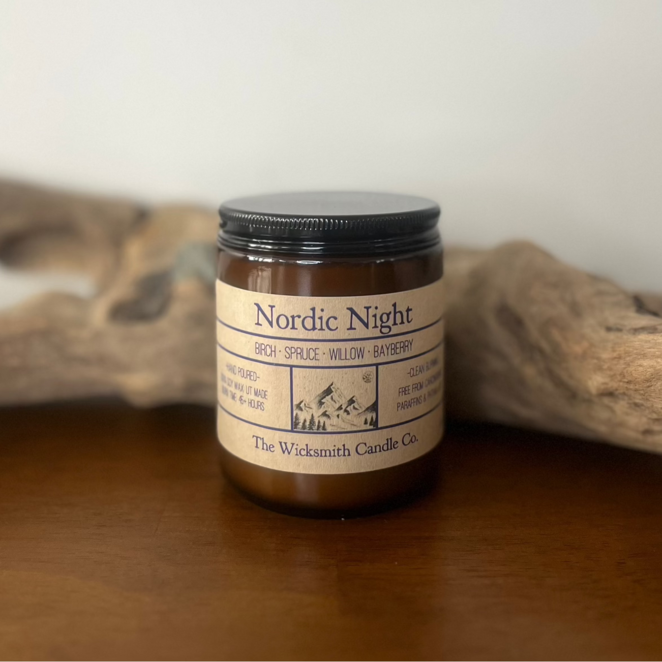 Amber glass candle jar labeled ‘Nordic Night’ by The Wicksmith Candle Co., featuring scent notes of birch, spruce, willow, and bayberry, placed on a wooden surface with a piece of driftwood in the background