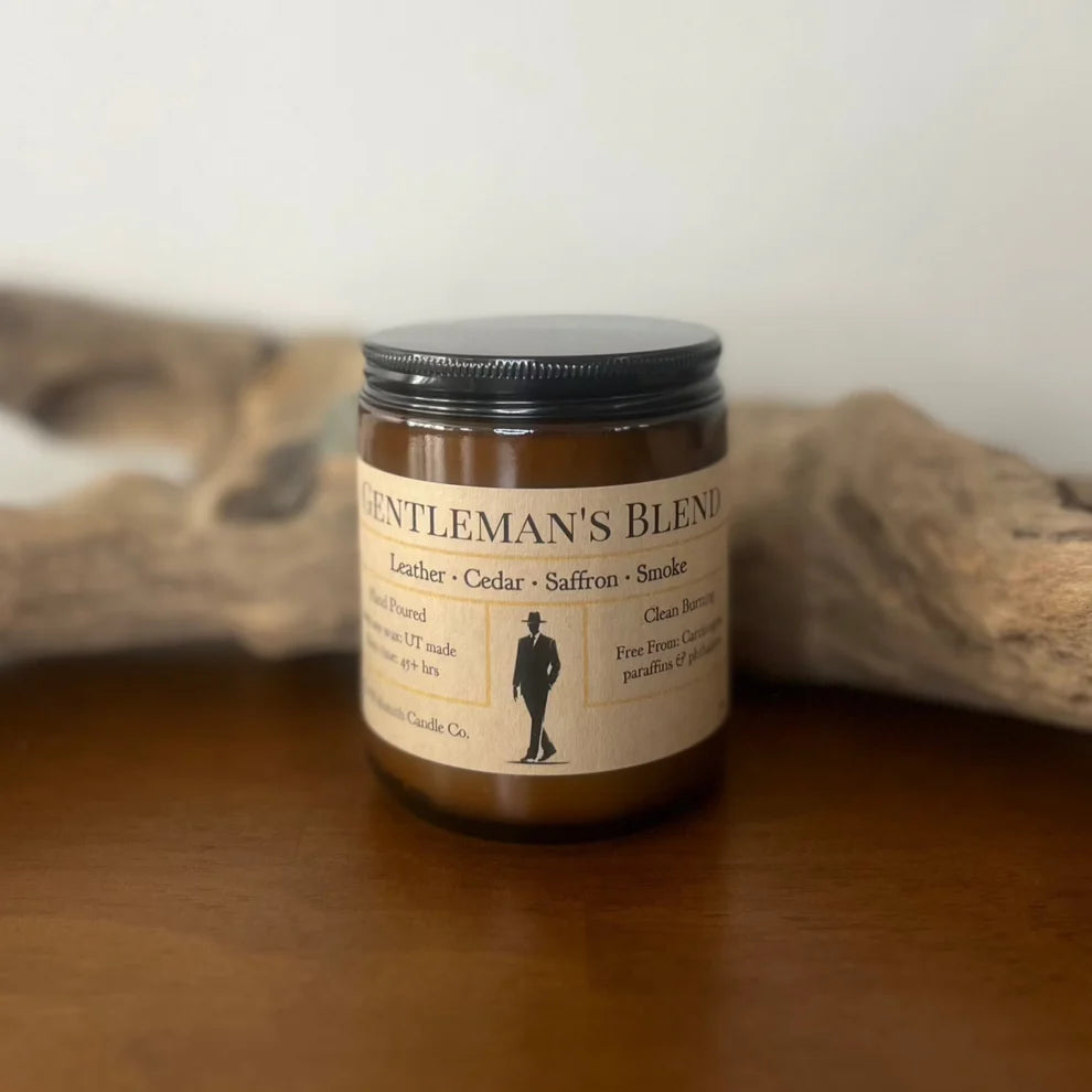 A brown glass jar candle labeled “Gentleman’s Blend” sits on a wooden surface with driftwood in the background. The label lists notes of leather, cedar, saffron, and smoke, and mentions the candle is hand-poured, clean-burning, and free from paraffins and phthalates.