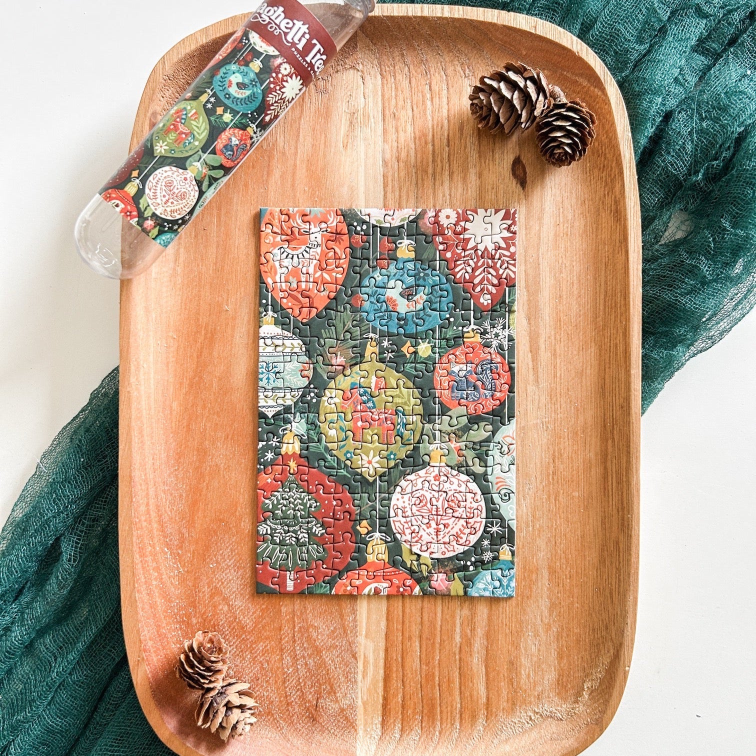 Finished mini ornament puzzle against a wooden tray with a dark green table runner, pinecones, and the empty tube.