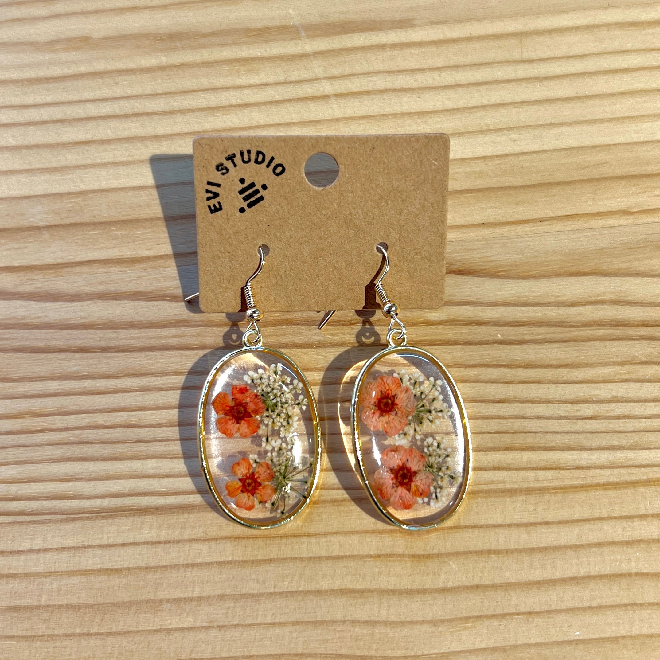 Gold rimmed large oval resin earrings with two orange pressed flowers and lots of small white flowers. Pictured on a wooden back ground. 