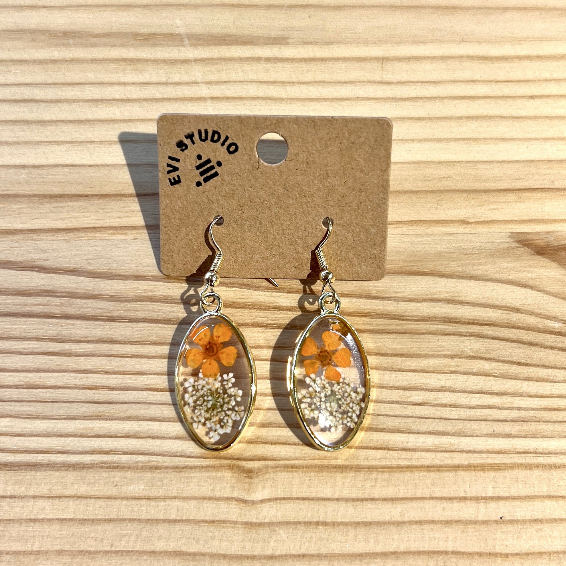 Gold rimmed oval resin earrings with pressed orange flower and small white flowers. Pictured on a wooden background. 