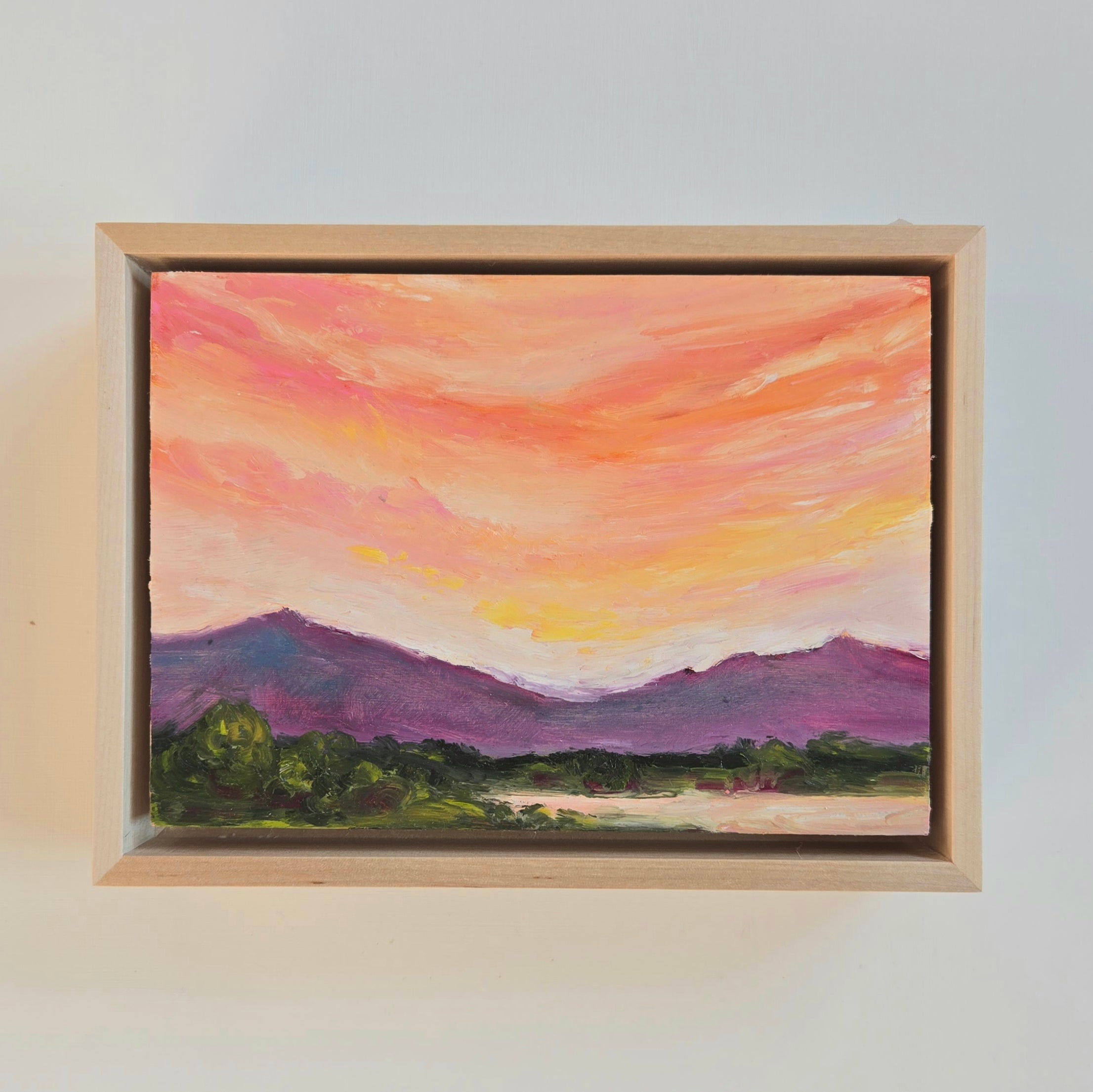 A pink, orange, and yellow sunset over a purple mountain range with green trees in the foreground. 