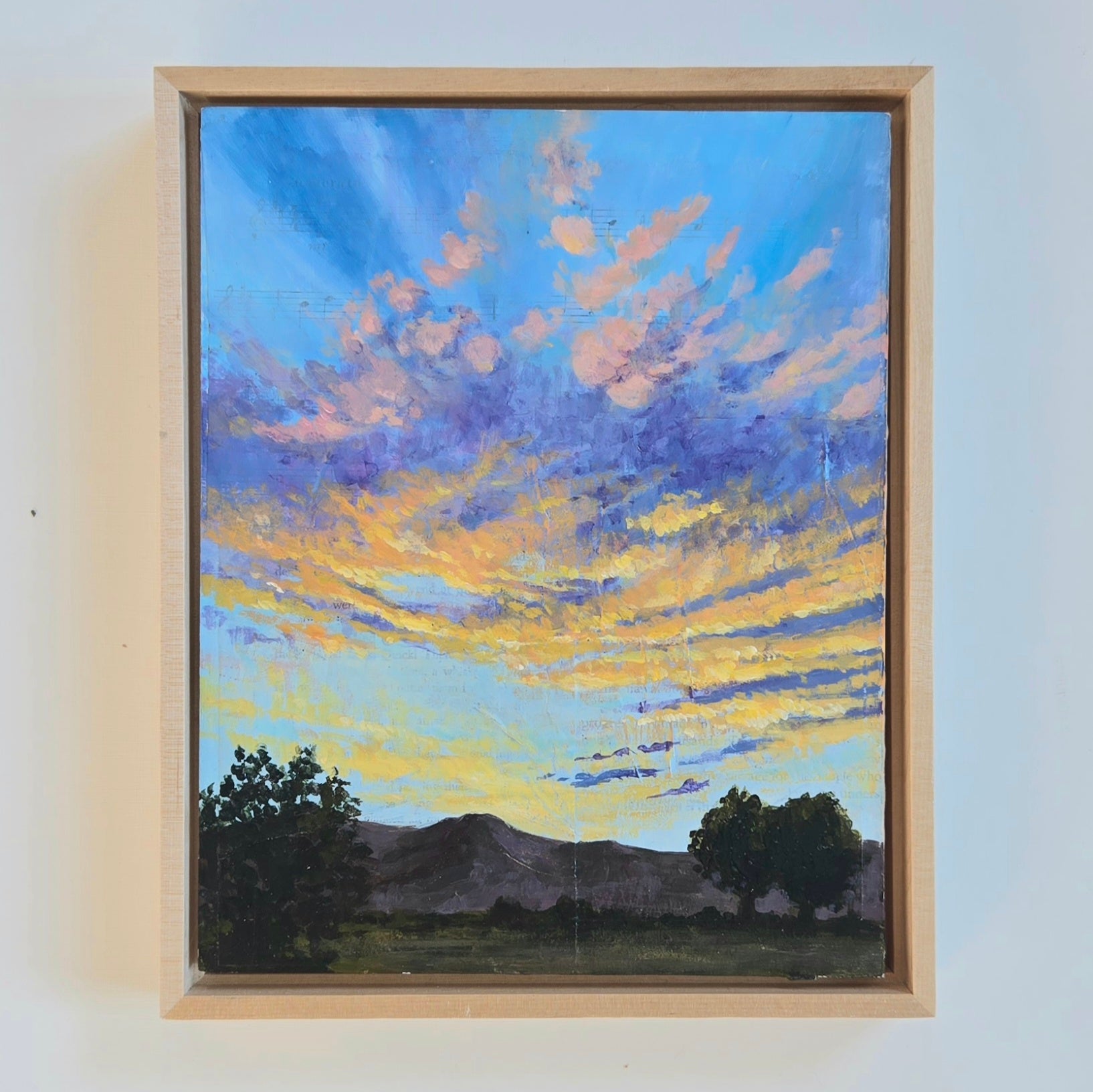 A bright blue sky at dusk is scattered with yellow, orange, and pink clouds. Below is a dark mountainscape with full trees and green plains. 