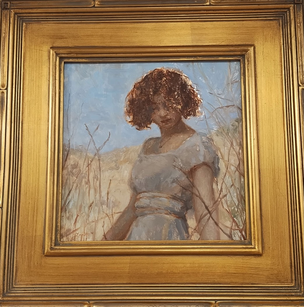 A brunette young woman looks down as she is wearing a blue-grey dress with short sleeves. You only see her from the waist up and her hair covers most of her face. She is surrounded by tall golden grasses and behind her is a pale blue sky. 