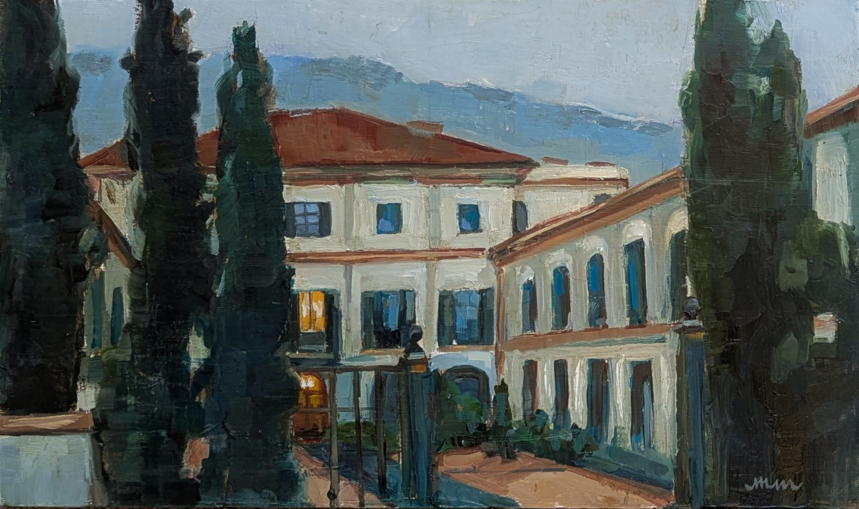 A building made of stucco with many balconies and windows and a red roof at dusk. The sky is pale and dark blue and you can see a light glowing from the main entrance of the building. There is an open cast iron gate in front and tall trees surround the gate. 