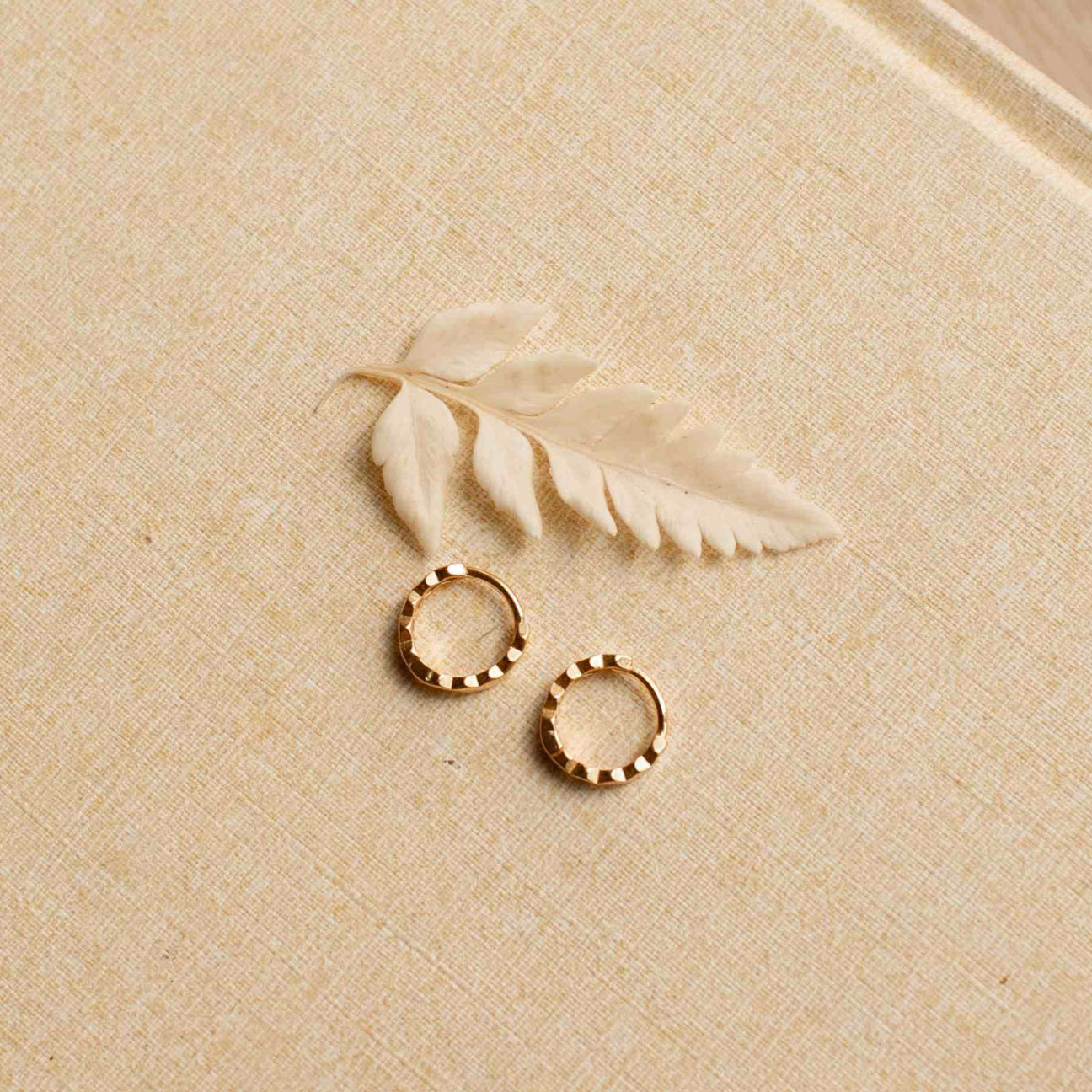 Two honeycomb huggie earrings laying flat on a linen surface. Next to them is a white leaf. 