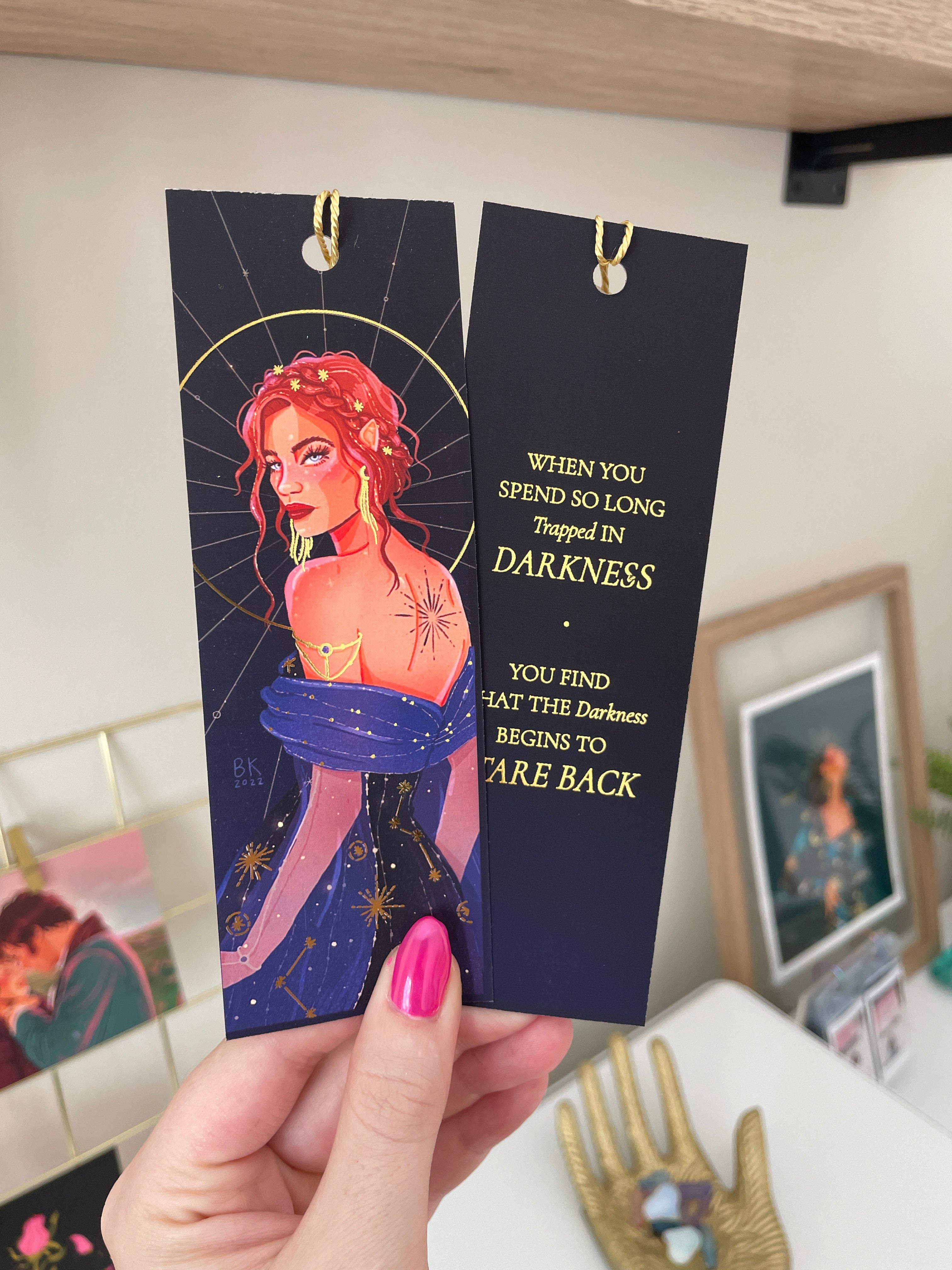 Nesta Archeron bookmark with an image of Nesta on one side and a gold foiled quote on the backside. Nesta has brown-red hair braided in an updo and she is looking over her shoulder at you. On her back is a tattoo of an exploding star and she is wearing a deep blue dress adorned with constellations. The back of the bookmark says, "When you spend so long trapped in darkness you find that the darkness begins to care back"