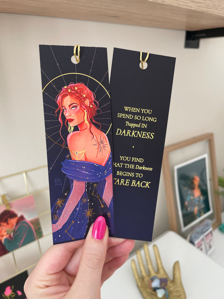 Nesta Archeron bookmark with an image of Nesta on one side and a gold foiled quote on the backside. Nesta has brown-red hair braided in an updo and she is looking over her shoulder at you. On her back is a tattoo of an exploding star and she is wearing a deep blue dress adorned with constellations. The back of the bookmark says, "When you spend so long trapped in darkness you find that the darkness begins to care back"