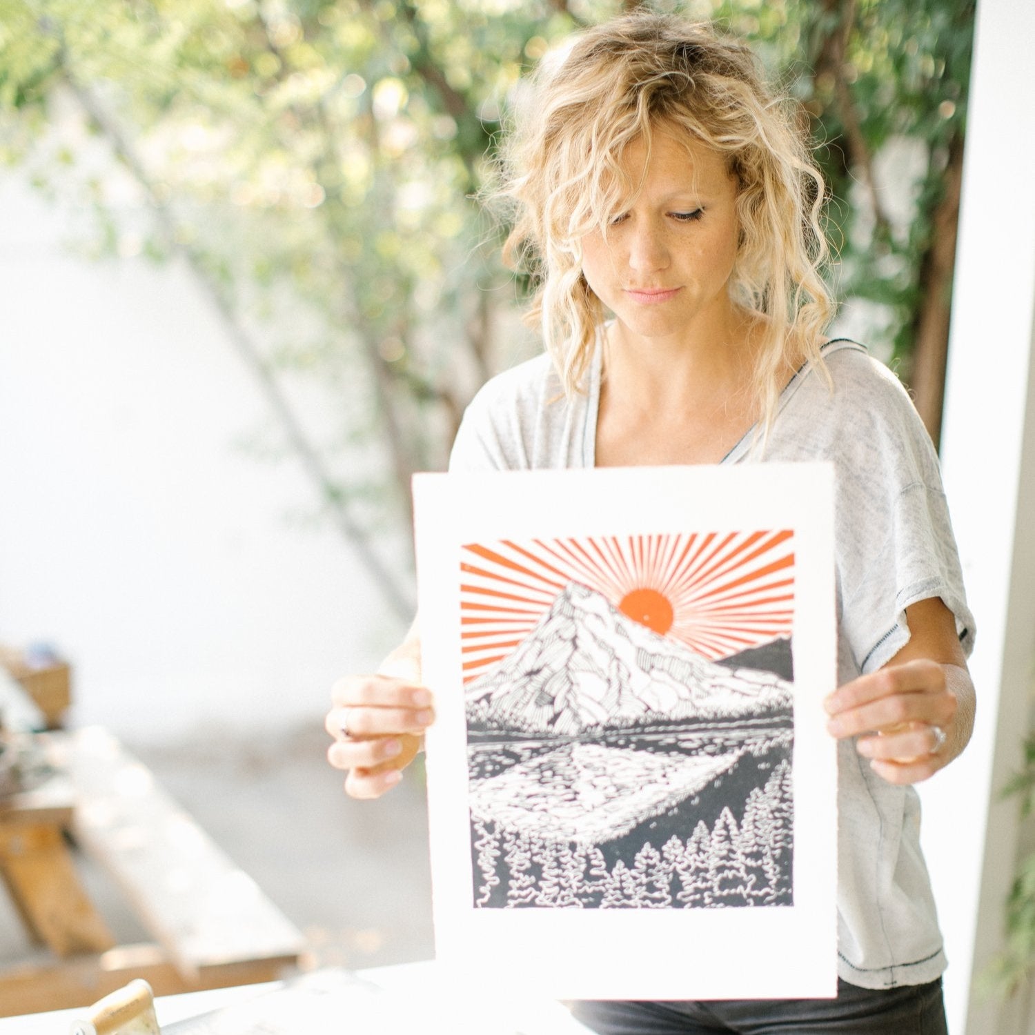 woman holding a hand carved and pressed black and white 11x14 print of a mountain range and pine trees with a red/orange sun shining across the sky