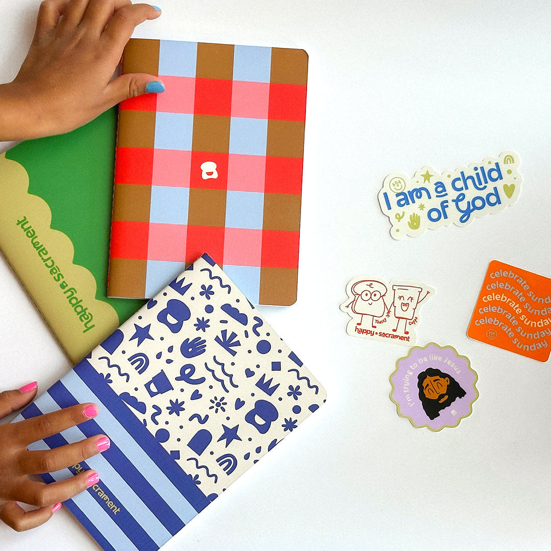 A collection of colorful notebooks with plaid and illustrated covers is arranged on a white surface, with hands holding two of the notebooks. Several playful Christian-themed stickers, including “I am a child of God” and “celebrate Sunday,” are displayed alongside them. 