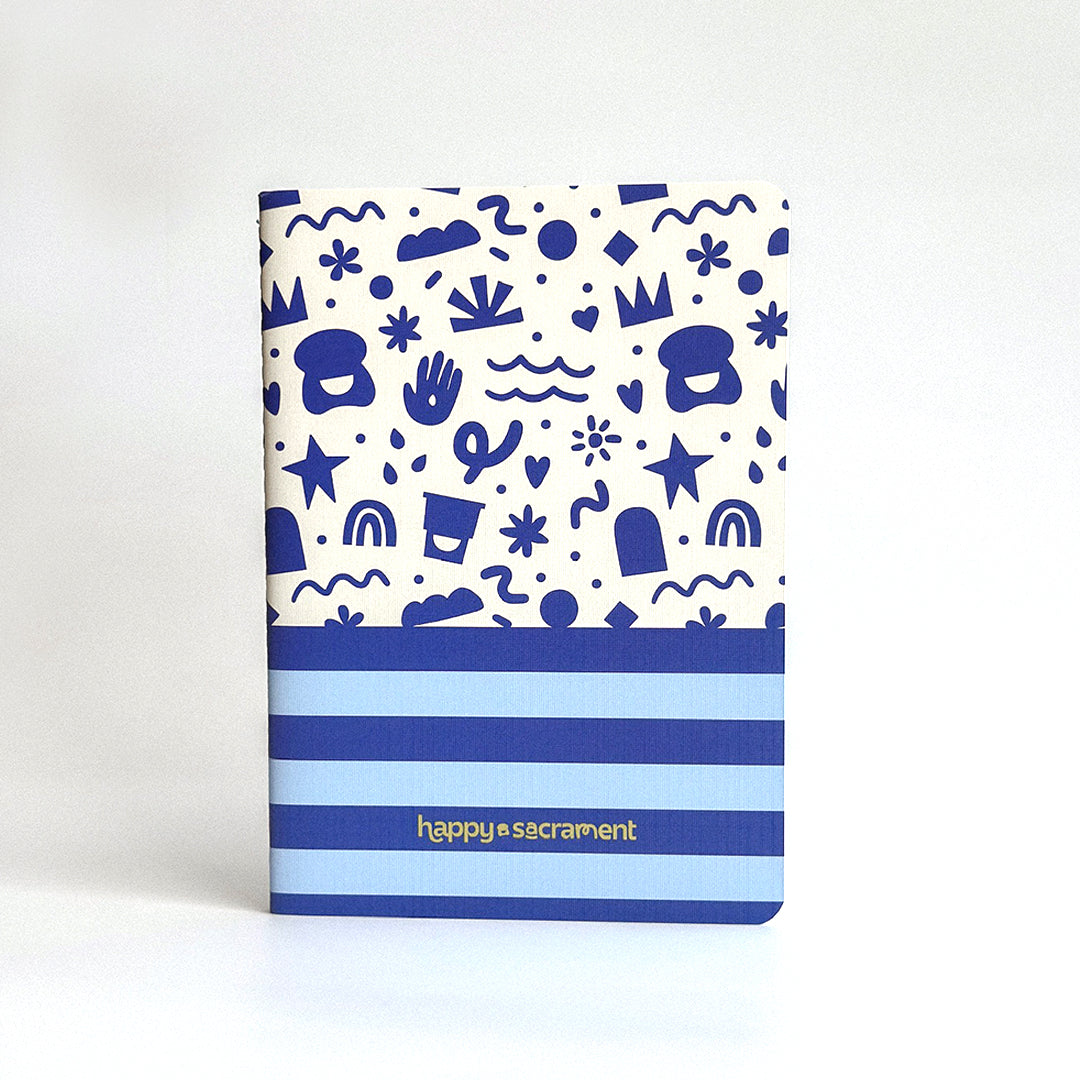 A cream-colored notebook features a playful blue pattern of crowns, hearts, stars, rainbows, and abstract shapes on the upper half. Blue horizontal stripes and a “happy sacrament” logo decorate the lower portion of the cover. 