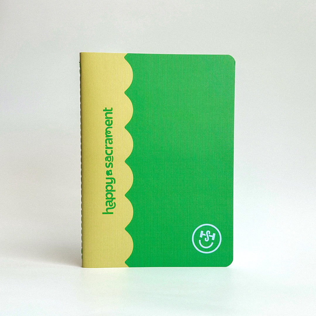 A green and yellow notebook with rounded corners is displayed against a light background, featuring a scalloped color-blocked design. The cover includes “happy sacrament” text and a white circular smiley logo.