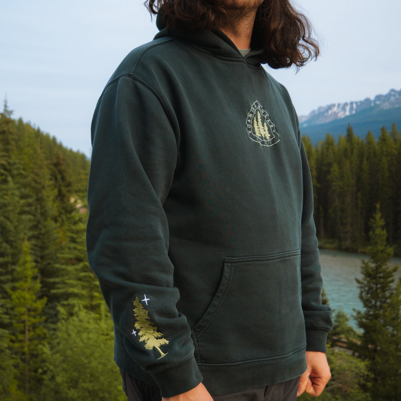 Image shows dark green sweatshirt worn by a man standing amongst pine trees. Hoodie has embroidered pine trees on chest and lower sleeve. 