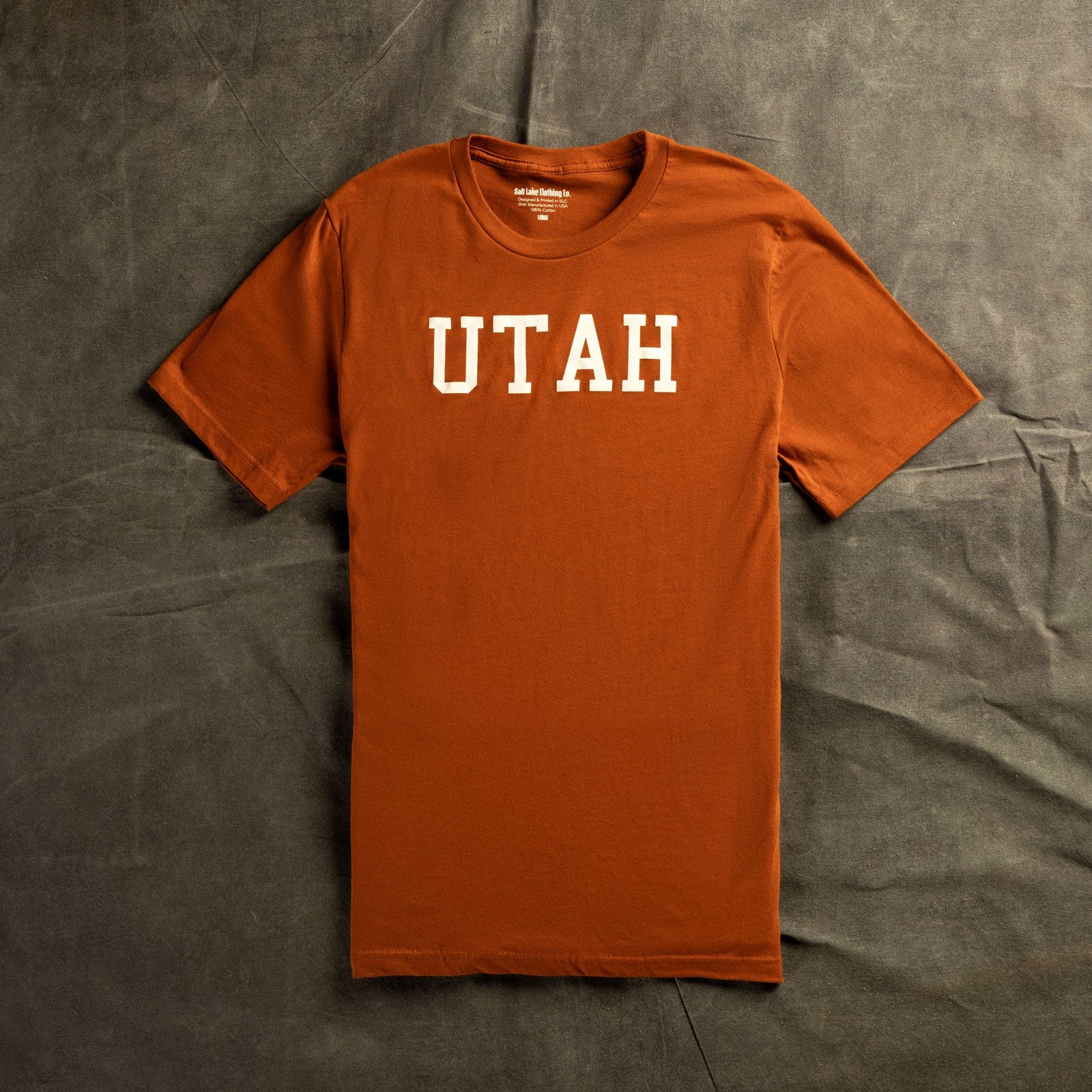 A rust orange tee shirt that says "UTAH" on the chest lying flat against a grey marble background