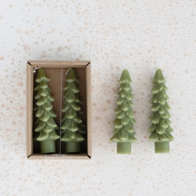 Two green tree-shaped candles on a textured white surface with one candle in an open box.
