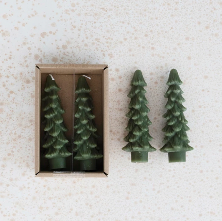 Two green tree-shaped candles on a textured beige surface with one candle in an open box.