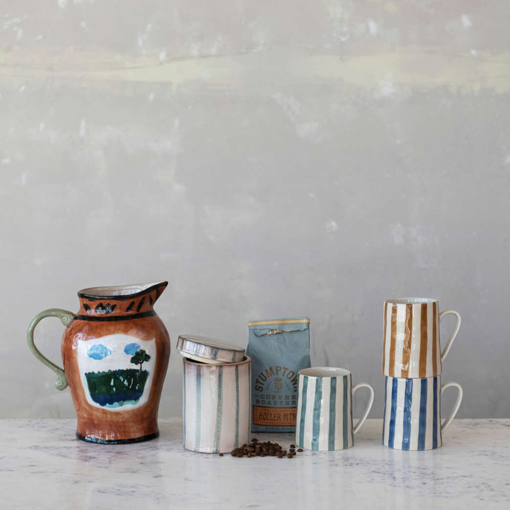 Set of ceramic items including a pitcher, canister, and mugs on a marble surface with a gray textured wall background.