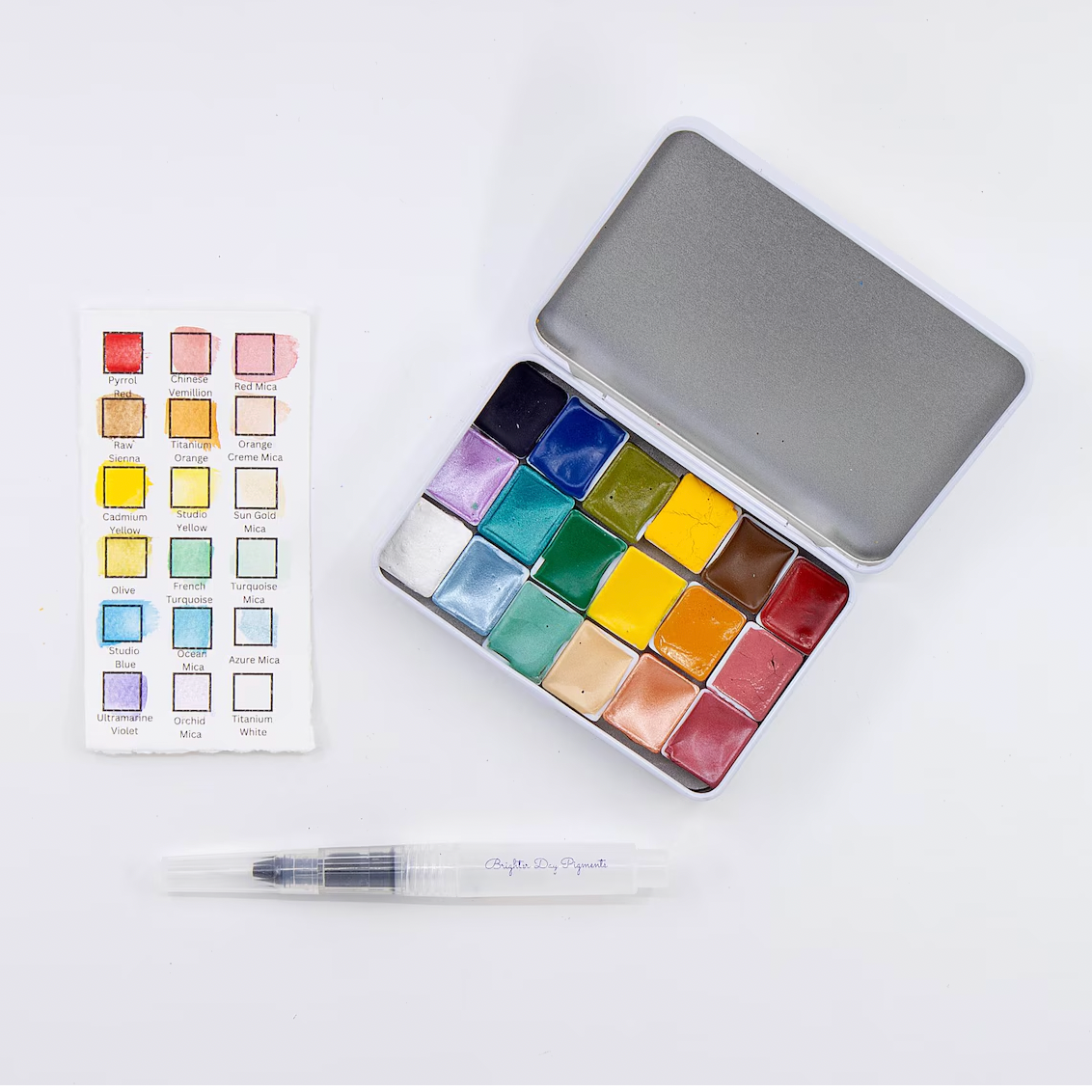 Set of watercolor paints in a tin with a color swatch and pen on a white background