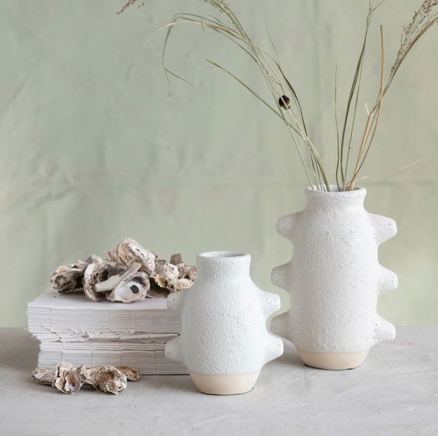 Two textured white vases with dried plants on a light surface and green background