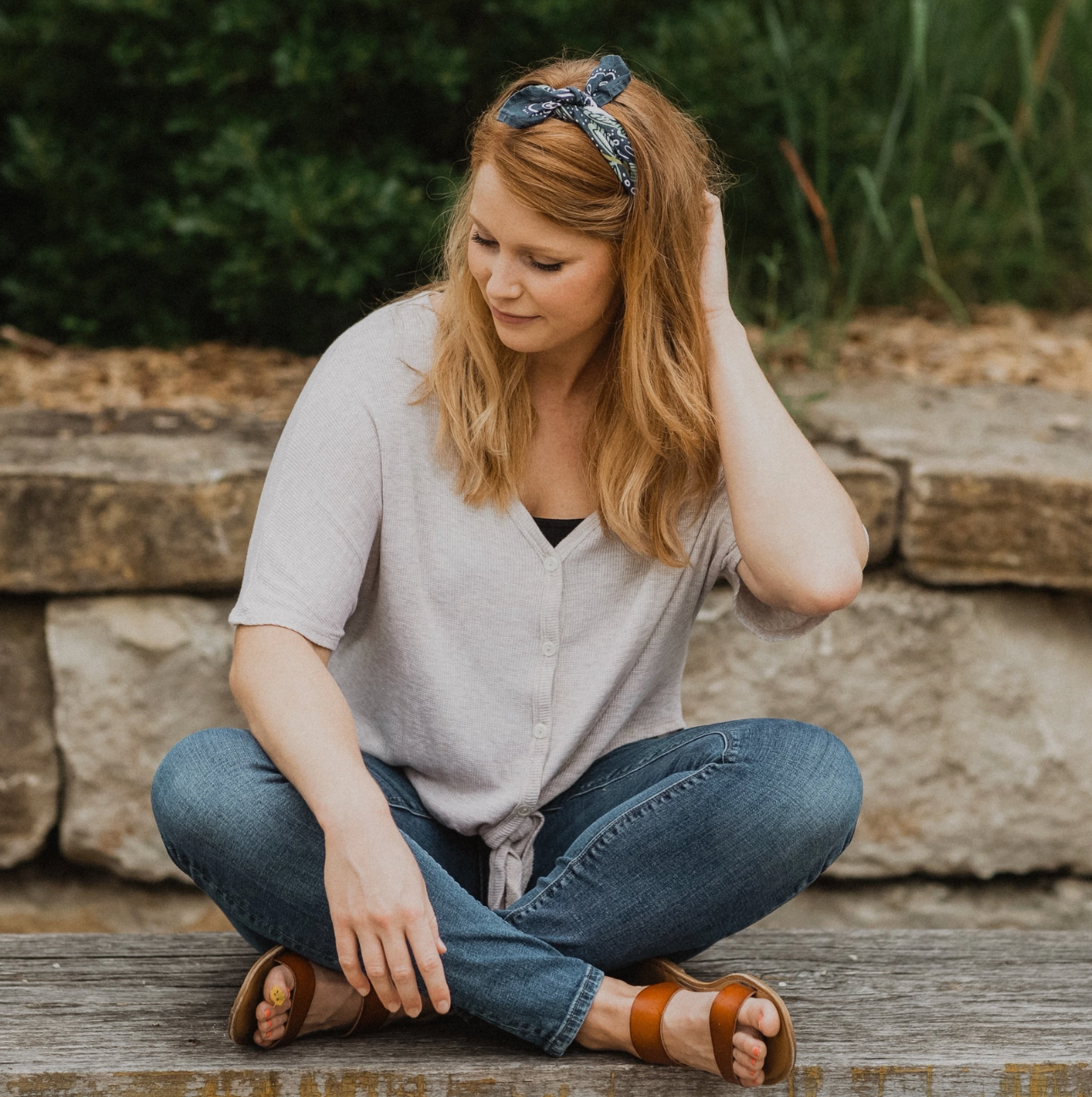 A woman with blonde hair wearing a blue patterned headband, a light gray tie-front short-sleeve top, blue jeans, and brown sandals, sitting cross-legged on a wooden surface in front of a stone wall and green foliage.
