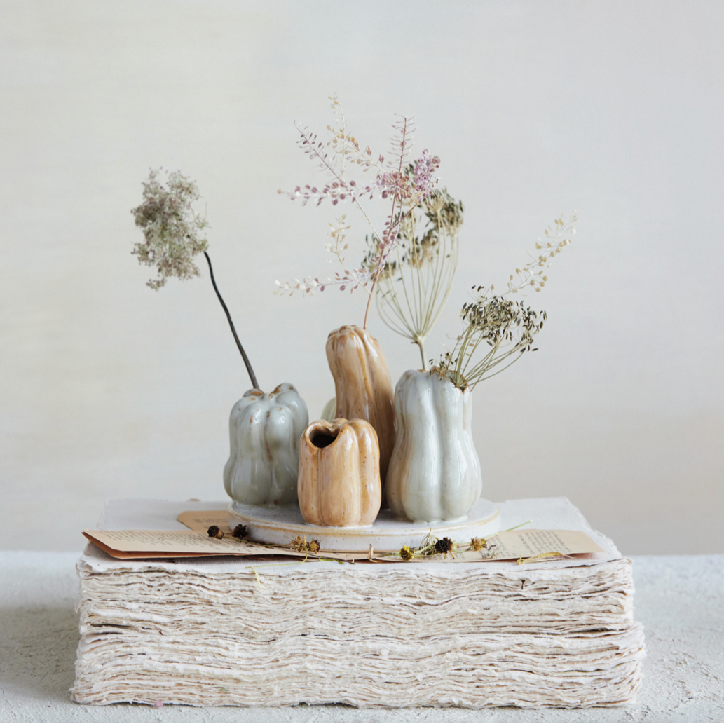 Four small ceramic vases in muted sage, cream, and natural wood tones, arranged on a stack of textured paper with dried flowers and small beads against a soft neutral background.