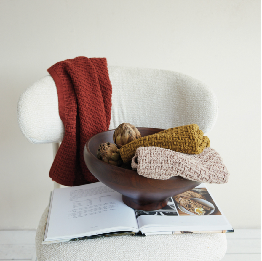 Three knitted tea towel in terracotta, mustard yellow, and light pink are styled on a white armchair with a dark wood bowl, artichokes, and an open magazine.
