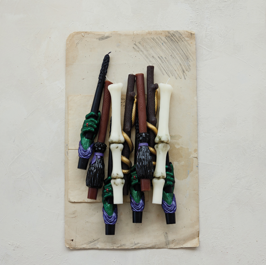 A collection of decorative witch-style candles arranged on a square piece of aged beige paper, photographed from above against a light textured wall.