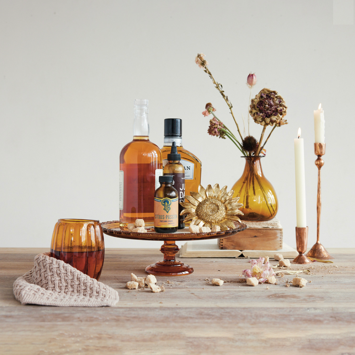 A styled tabletop still life with amber glass bottles, a small amber tumbler, a candlestick holder with a white candle, dried flowers in a glass vase, books, and a beige knit cloth on a wooden surface against a plain light background.