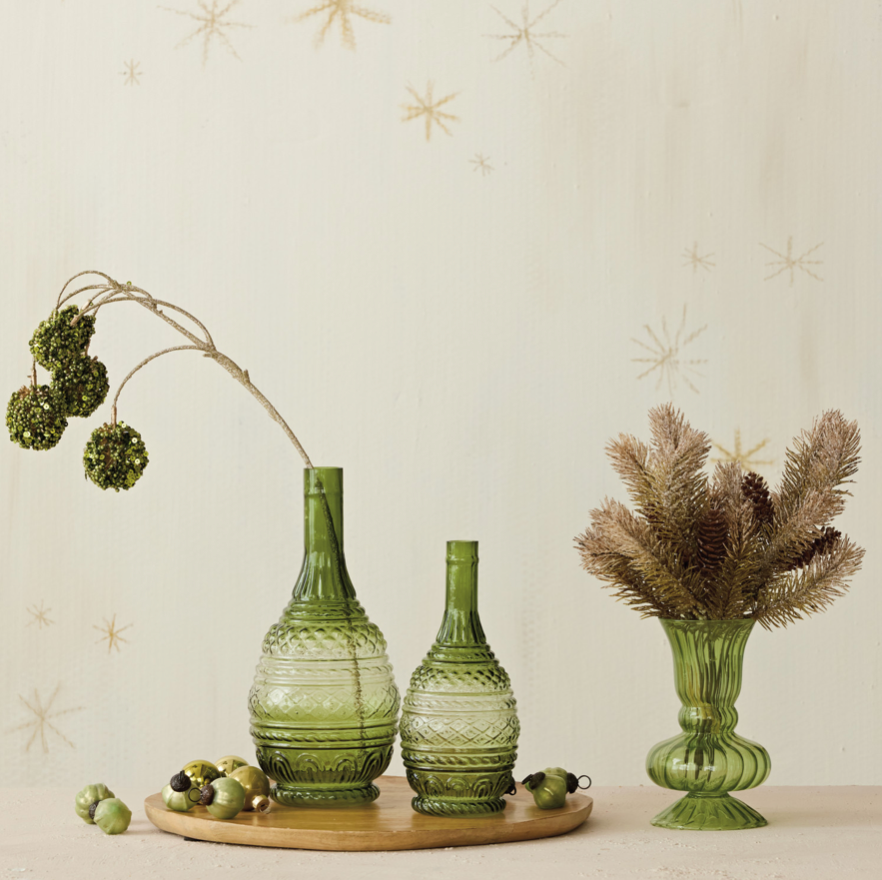 Three green glass vases of different shapes and sizes arranged on a round wooden tray, with dried greenery and pine branches placed in them, against a light cream wall with subtle star decals.