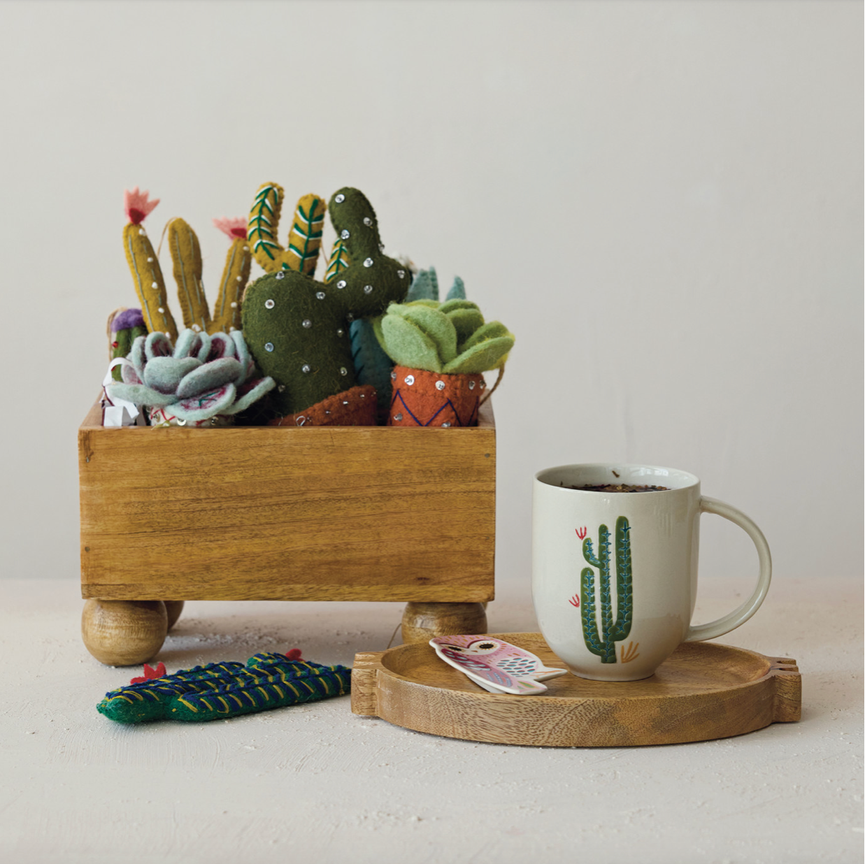 A wooden planter box filled with colorful felt succulents and cacti, styled on a white surface next to a ceramic mug with a cactus print on a wooden tray.