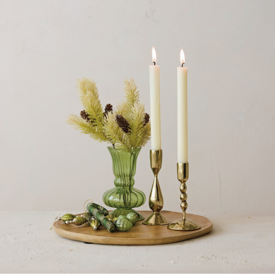 A styled tabletop arrangement on a round wooden tray, featuring two lit white taper candles in brass candlesticks, a small green ribbed glass vase with pine branches, and green glass ornaments against a plain off-white wall.