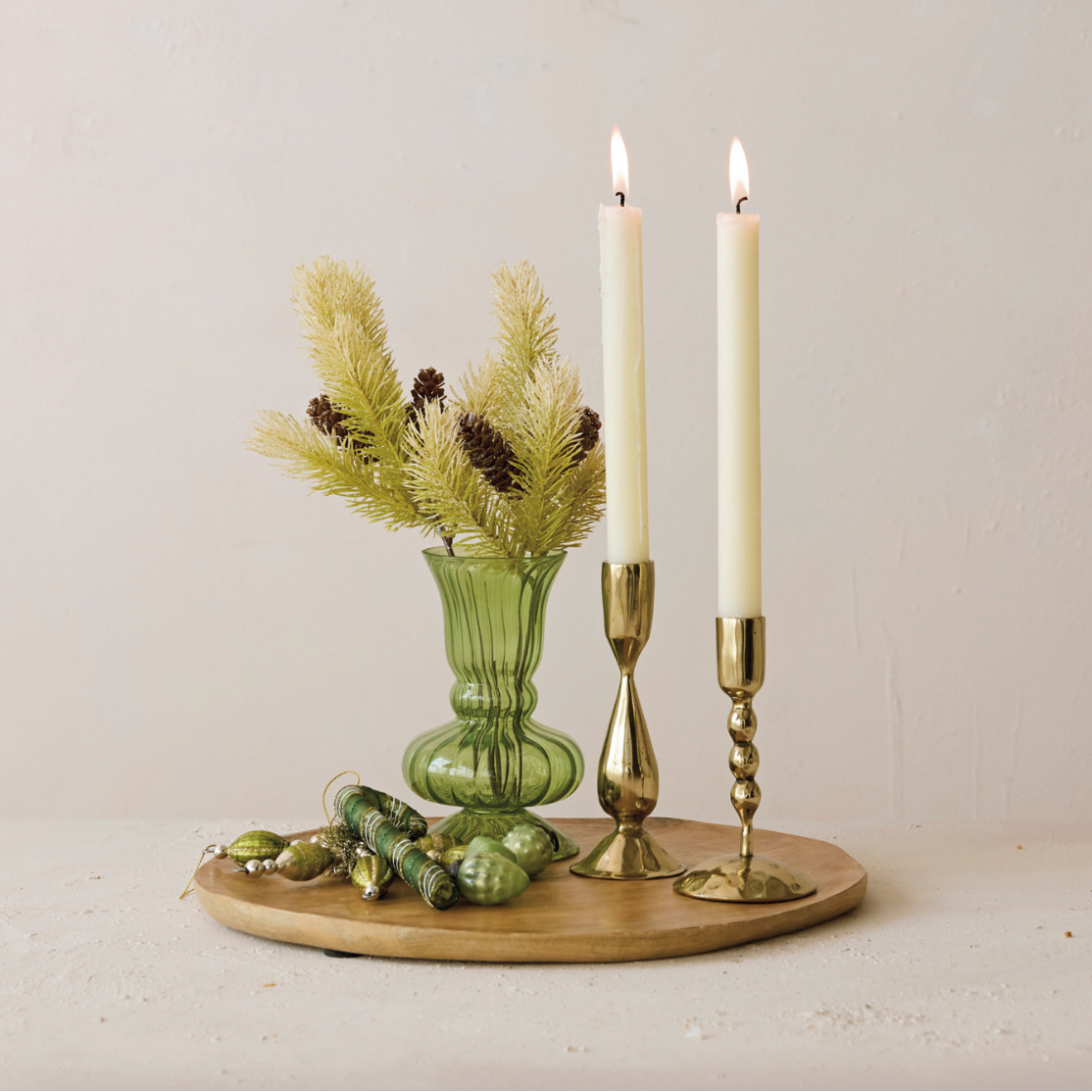 A styled tabletop arrangement on a round wooden tray, featuring two lit cream taper candles in brass holders, a small green ribbed glass vase with pine branches and pinecones, and several green decorative ornaments against a plain light beige wall.