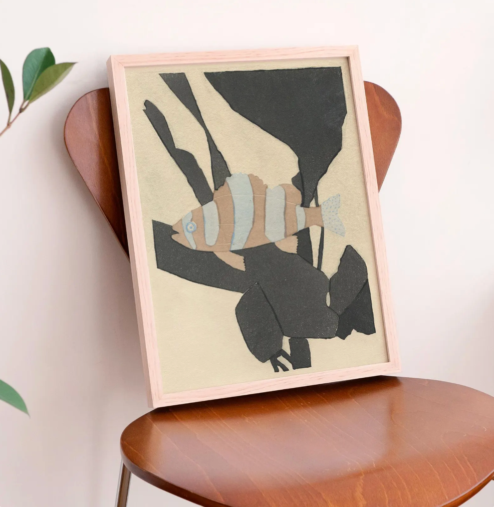 Framed abstract fish wall art leaning on a wooden chair against a light wall, with a plant leaf visible at the left edge.