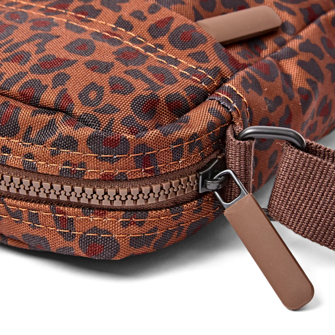 Close-up of a brown and black leopard-print crossbody bag with a brown zipper pull and matching fabric strap, shown on a white background.
