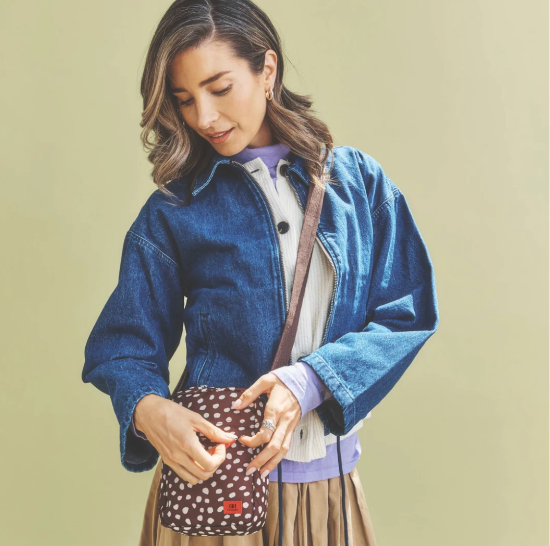 A woman wearing a blue denim jacket and white sweater looks down at a brown polka-dot crossbody bag with white dots and a brown strap, set against a solid light olive-green background.