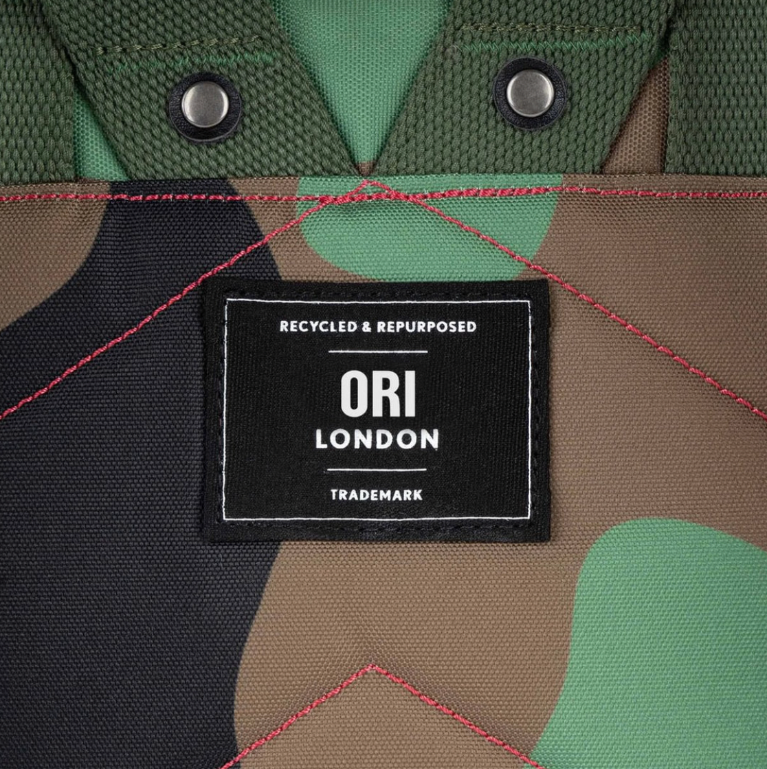 Close-up of a camouflage fabric product with green, brown, and black patches, red stitching, metal grommets, and a central black label reading 'RECYCLED & REPURPOSED ORI LONDON TRADEMARK'.
