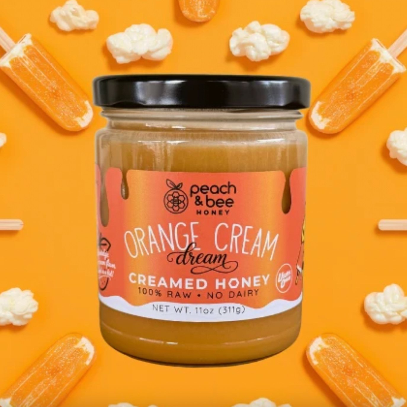 A jar of Peach & Bee Honey 'Orange Cream Dream' creamed honey centered on a bright orange background decorated with popsicle sticks and popcorn. The jar has a black lid and a peach-to-orange label.