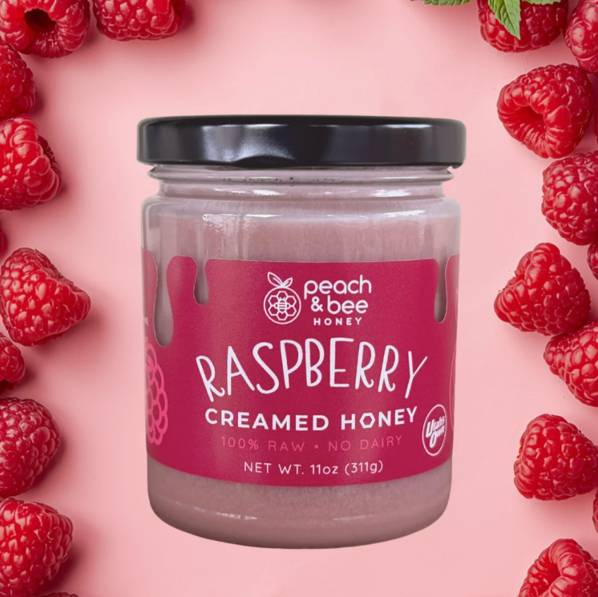 A jar of Peach & Bee Honey Raspberry Creamed Honey centered on a pink background, surrounded by fresh raspberries. The jar has a black lid and a pink label with white text.