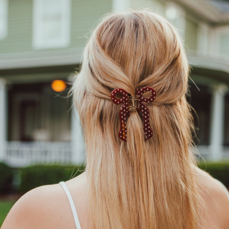 A blonde woman viewed from behind, wearing a brown & white polka dot bow-shaped hair clip in her low ponytail, with a blurred house porch in the background.
