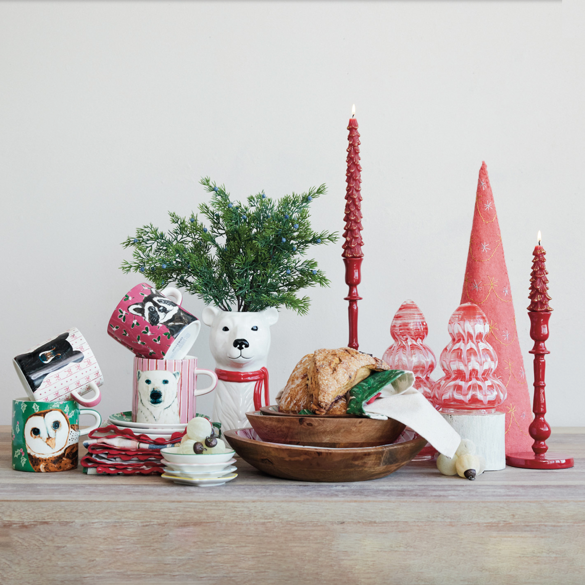 A styled holiday tabletop arrangement featuring multiple ceramic mugs, red taper candles, pink Christmas tree ornaments, a small green plant in a bear-shaped holder, and a wooden bowl with bread and greenery, set against a plain light background.