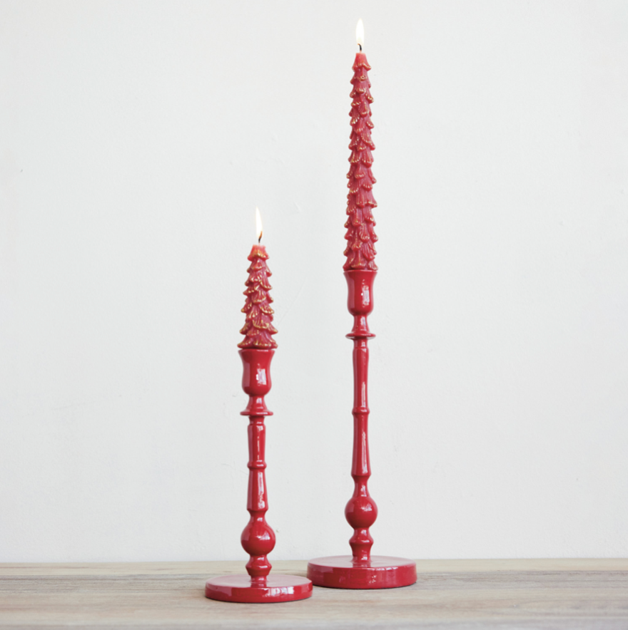 Two red taper candles with ornate, textured tree-like tops and turned wooden bases, standing on a light wood surface against a plain off-white wall.
