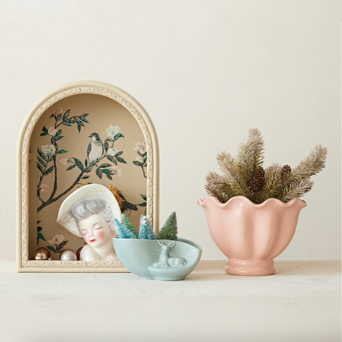 A styled tabletop arrangement on a light neutral background featuring a pink scalloped ceramic planter with pine branches, a pale blue bowl with miniature trees, and an arched cream frame containing floral artwork and a porcelain woman's face.