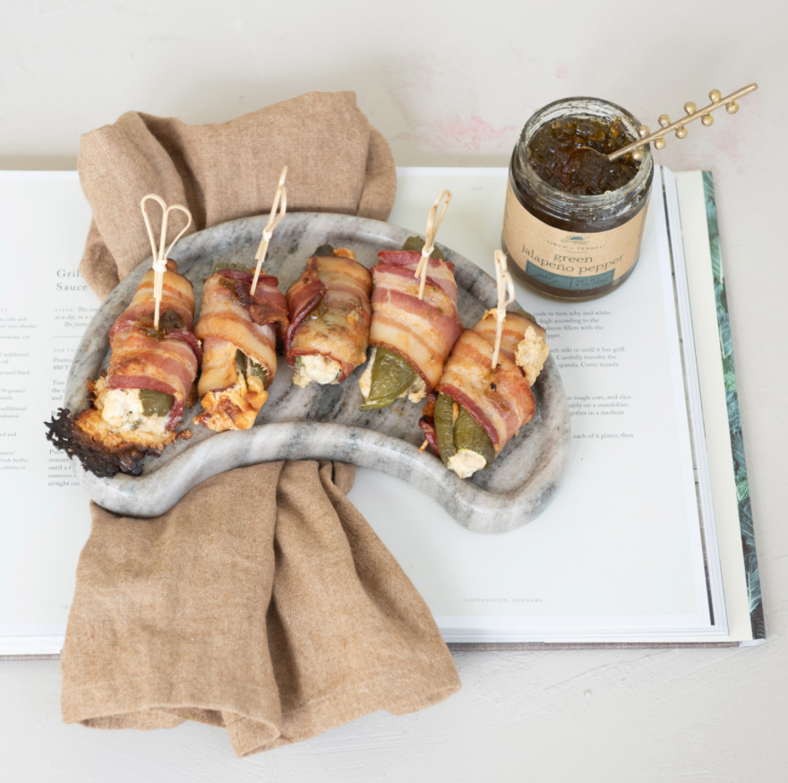 Five bacon-wrapped stuffed grape leaves on a gray marble serving platter, styled on an open cookbook with a beige linen napkin and a small jar of herb spread nearby.
