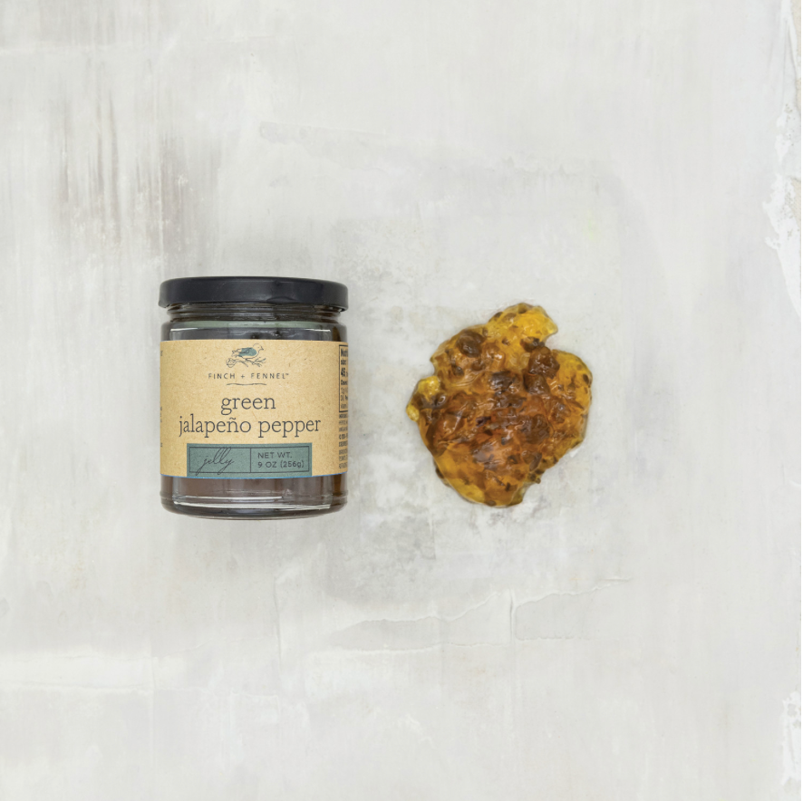 A glass jar of Fresh & green jalapeño pepper with a black lid and beige label, placed on a textured off-white surface next to a whole cooked jalapeño pepper.