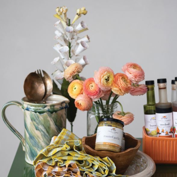 A styled tabletop arrangement featuring a jar of roasted garlic, peach ranunculus flowers in a glass vase, a green marbled pitcher with wooden spoons, a yellow-and-white checkered cloth, and several bottles in an orange basket against a plain light gray background.