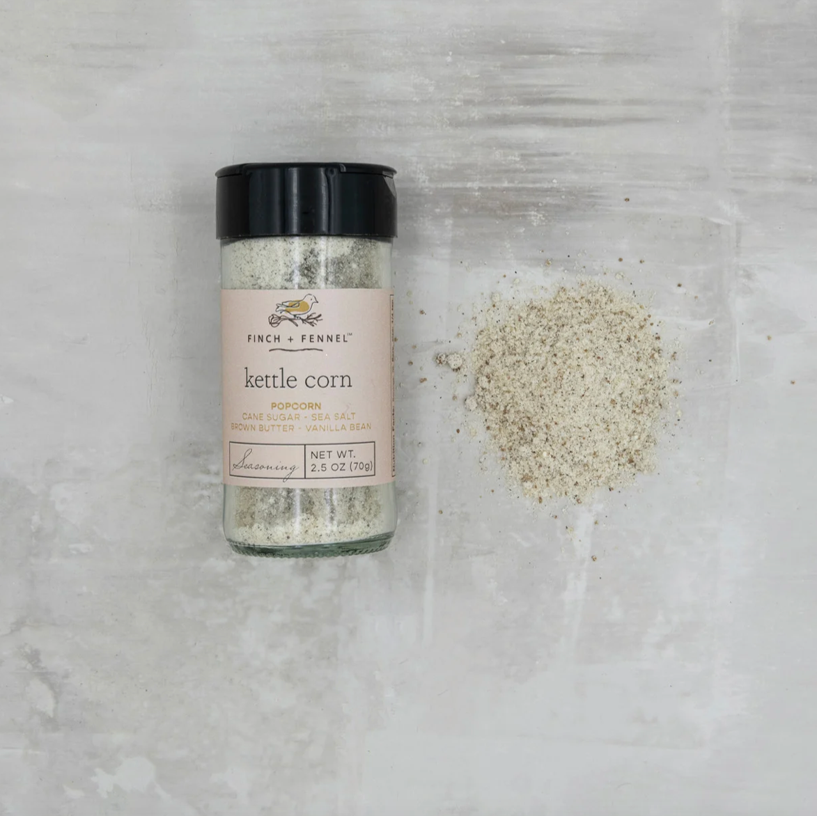 A bottle of Finch & Ferrie Kettle Corn seasoning with a black cap and a pink label, placed on a light gray textured surface next to a small pile of the seasoning.
