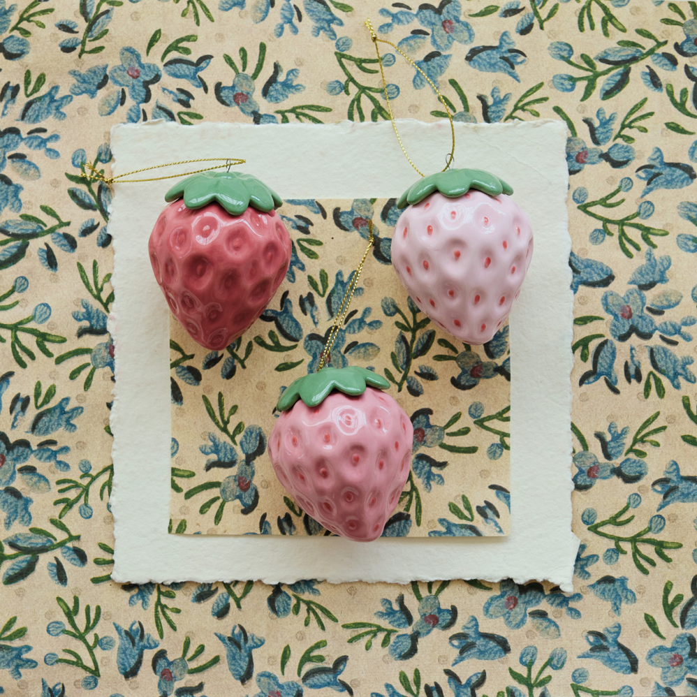 Three decorative strawberry ornaments in pink shades with green tops, hanging from a gold string against a layered floral paper background.