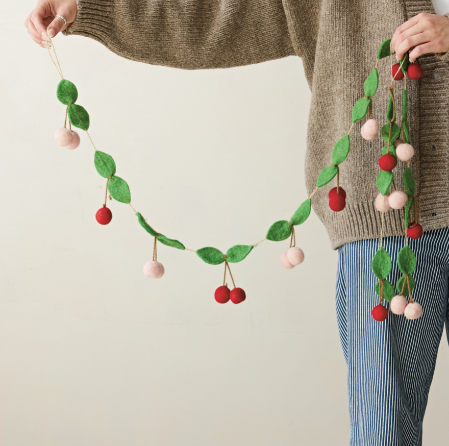 A person holds up a felt cherry garland with green leaves, red cherries, and pale pink pom-poms against a plain light beige background.