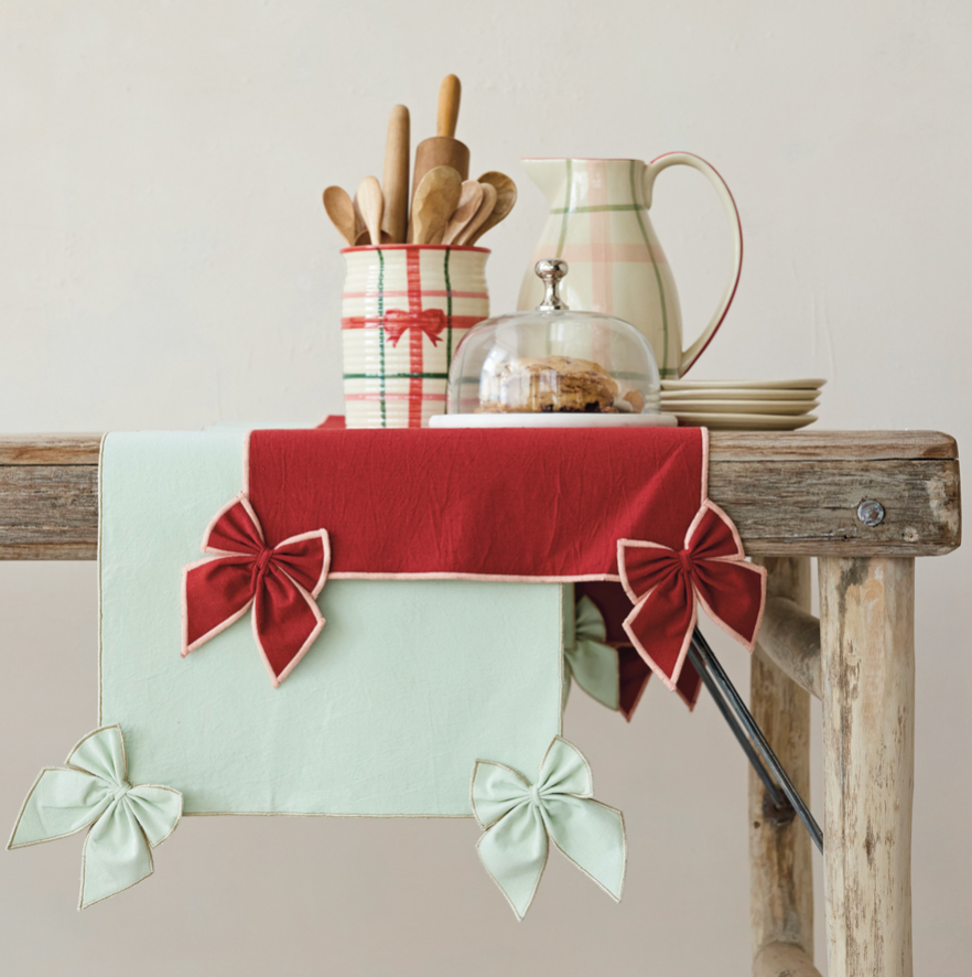 A festive holiday table runner with a red top band, a pale mint green main body, and matching red and mint green bow details, styled on a rustic wooden table with kitchen utensils and a cream pitcher.